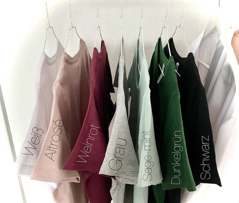 Pode incluir: Oito t-shirts penduradas em cabides, mostrando diferentes cores: branco, rosa claro, bord&ocirc;, cinza, verde claro, verde escuro, preto. As t-shirts s&atilde;o etiquetadas com as seguintes cores: Wei&szlig;, Altros&eacute;, Weinrot, Grau, Sage-mint, Dunkelgr&uuml;n, Schwarz.