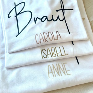 Pode incluir: Quatro camisetas brancas empilhadas umas sobre as outras. A camiseta de cima tem a palavra "Braut" escrita em caligrafia preta. A segunda camiseta tem o nome "Carola" escrito em ouro rosa. A terceira camiseta tem o nome "Isabell" escrito em ouro. A camiseta de baixo tem o nome "Anne" escrito em ouro.