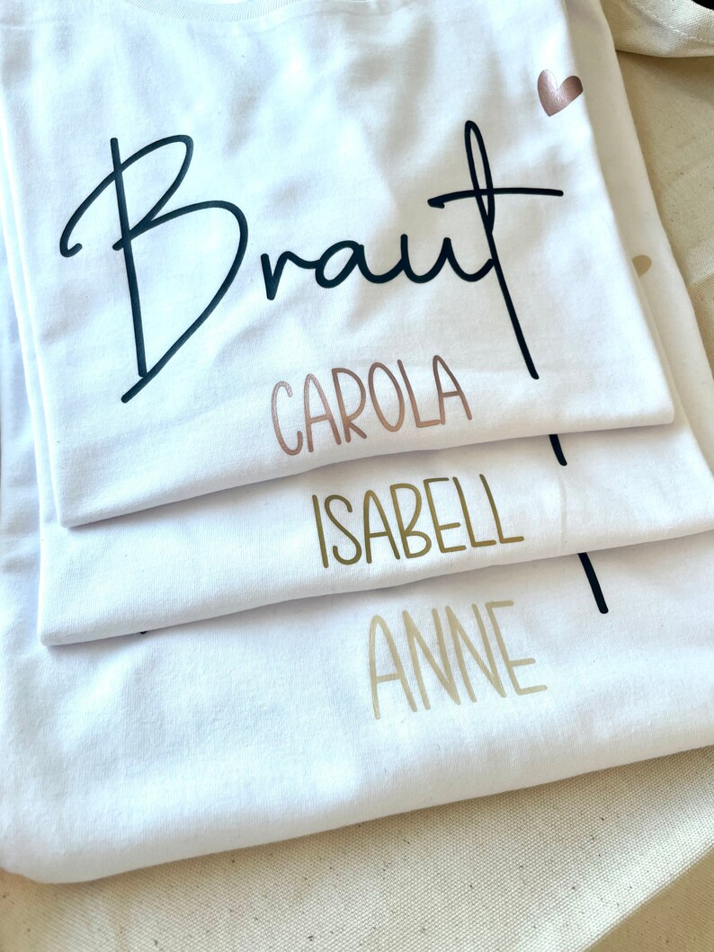 Pode incluir: Quatro camisetas brancas empilhadas umas sobre as outras. A camiseta de cima tem a palavra "Braut" escrita em caligrafia preta. A segunda camiseta tem o nome "Carola" escrito em ouro rosa. A terceira camiseta tem o nome "Isabell" escrito em ouro. A camiseta de baixo tem o nome "Anne" escrito em ouro.