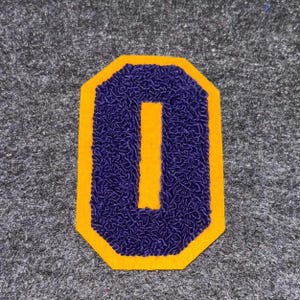 May include: A purple letter "I" patch with a gold border and a yellow outline. The letter is embroidered with a textured design. The patch is a classic design, suitable for jackets, bags, or other apparel.