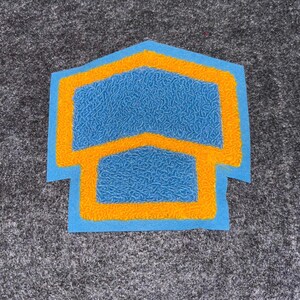May include: A blue and orange patch with a textured surface. The patch has a house-like shape with a blue interior and orange outline. The patch is on a gray surface.
