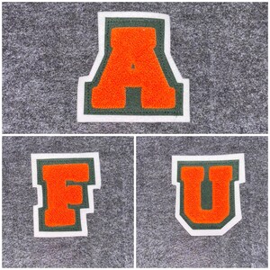 May include: Three felt letter patches: an orange "A", "F", and "U". Each letter has a white border, a green outline, and a textured orange center. The patches are displayed on a gray felt background.