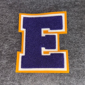 以下が含まれることがあります： 紫色の文字「E」のパッチ。表面はテクスチャ加工され、白で縁取られ、金色の縁で囲まれています。 パッチは、スタジアムジャンパーなどのアパレルのためである可能性が高く、灰色の背景に配置されています。
