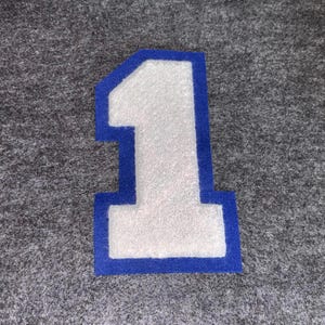 May include: A white felt number 1 patch with a blue border, set against a gray background. The number is a classic varsity style, suitable for jackets, bags, or other fabric items.