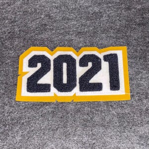 May include: A yellow, white, and navy blue felt patch with the number "2021" stitched in a varsity style. The patch has a white background with navy blue numbers and a yellow border. The patch is on a gray felt surface.