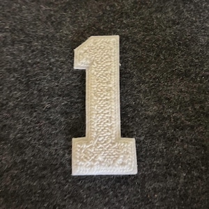 Puede incluir: Un número uno blanco y texturizado, probablemente un artículo decorativo o parte de un conjunto. El número tiene un patrón detallado y punteado en su superficie. El fondo es una tela gris oscuro texturizada.