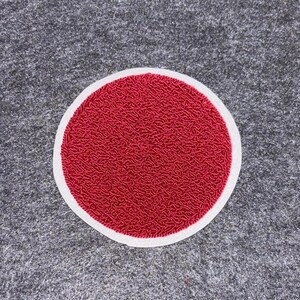 Puede incluir: Un parche redondo rojo con una superficie texturizada y un borde blanco. Se encuentra sobre un fondo gris texturizado. El rojo es vibrante y la textura parece estar hecha de pequeños hilos entrelazados. Diseño simple y llamativo.