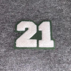 May include: A white, felt number 21 patch with a green border. The patch is displayed on a grey, textured background. The number is in a varsity style font.