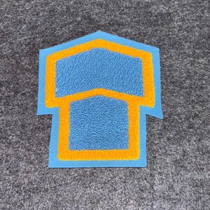 May include: A blue and yellow felt patch in the shape of a house. The patch has a textured blue interior with a yellow border and outline. The patch is on a gray surface.