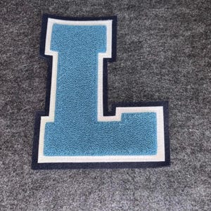 May include: A light blue felt letter "L" patch with a white border and navy blue trim, set against a gray background. The patch has a textured surface, commonly used for varsity jackets and apparel.