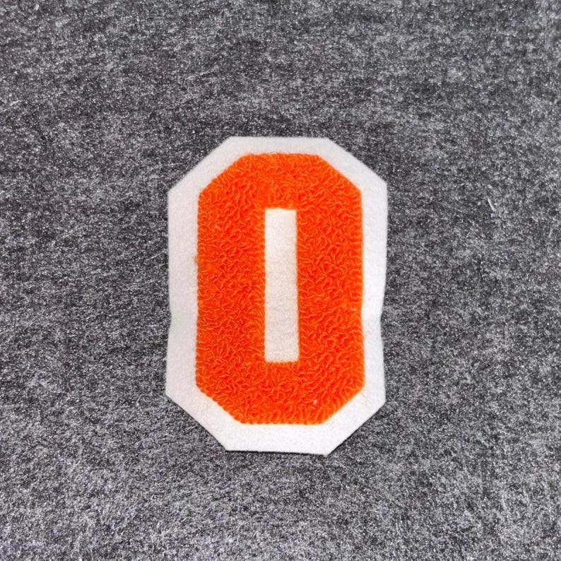 May include: An orange felt letter "O" patch with a white border. The letter has a textured appearance and is set against a gray felt background. This patch is suitable for jackets, bags, or other fabric items.