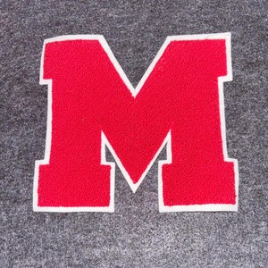 May include: A red letter "M" patch with a white border, set against a gray background. The letter has a textured appearance, suggesting a felt or embroidered material. This patch is suitable for jackets, bags, or other fabric items.