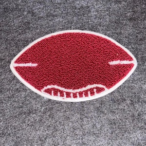 Pode incluir: Um patch vermelho em forma de bola de futebol com borda e detalhes brancos. O patch tem uma superfície texturizada e está sobre um fundo cinza. O patch é provavelmente para um item com tema esportivo.
