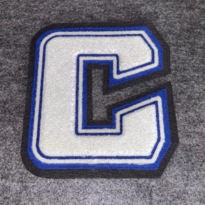May include: A white felt letter "G" patch with a blue and black border. The letter is outlined in blue stitching and has a black border. The patch is on a gray felt background.