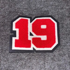 May include: A red and white number 19 patch with a black border. The number is in a varsity style font, with a textured appearance. The patch is likely for a jacket or other apparel.