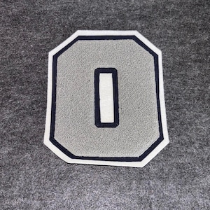 Puede incluir: Parche con la letra "O" gris texturizada, con borde blanco y contorno azul marino. El centro de la letra es blanco con un borde azul marino. Sobre un fondo gris.