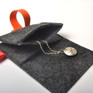 以下が含まれることがあります： 赤い「HAND MADE」のラベルとオレンジ色のループハンドルが付いたグレーのフェルトポーチ。ポーチが開いており、丸いペンダントが付いたシルバーネックレスが見える。