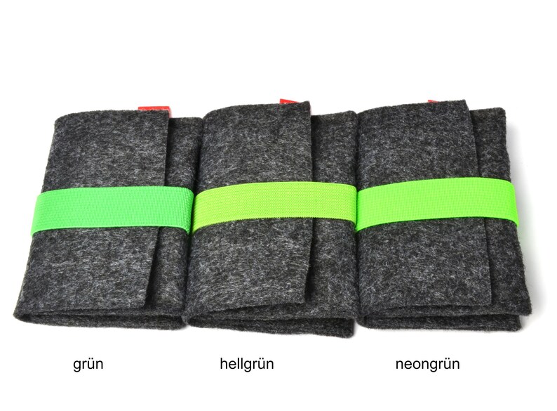 May include: Three rectangular dark gray felt cases, each secured with a bright neon green elastic band. The cases are arranged side-by-side, and each features a small red tag at the top edge. Below each case, the text reads gr&uuml;n, hellgr&uuml;n, and neongr&uuml;n.
