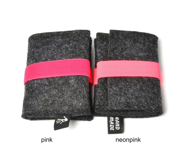 May include: Two rectangular dark gray felt cases secured with bright pink elastic bands. Each case features a small black fabric tag at the bottom edge. The left tag displays a white hand icon, while the right tag features the text HAND MADE in white.