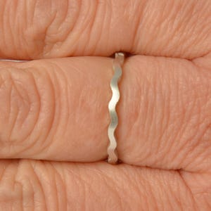 Könnte beinhalten: Ein silberner Ring mit einem wellenförmigen Design. Der Ring ist schmal und wird an einem Finger getragen. Das Design besteht aus einer Reihe verbundener Kurven, die ein strukturiertes Aussehen erzeugen. Es ist ein schlichtes, elegantes Schmuckstück.