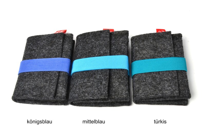 May include: Three rectangular dark gray felt cases, each secured with a horizontal elastic band. The bands are colored royal blue, medium blue, and turquoise from left to right. Each case features a small red fabric tag at the top edge.