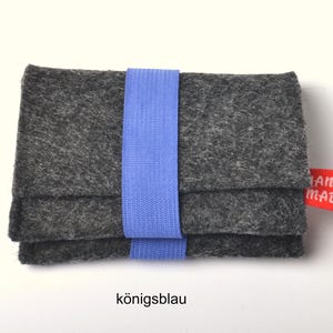 May include: A rectangular dark gray felt wallet secured with a light blue elastic band. The wallet features a small red fabric tag on the right side with the white text "HAND MADE" printed on it. The overall design is simple and functional.