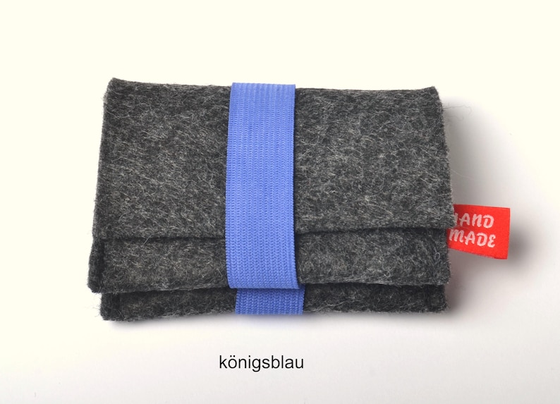 May include: A rectangular dark gray felt wallet secured with a light blue elastic band. The wallet features a small red fabric tag on the right side with the white text "HAND MADE" printed on it. The overall design is simple and functional.