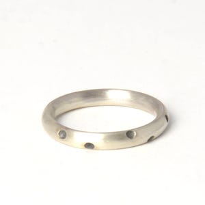 Könnte beinhalten: Ein silberner Ring mit einer Reihe kleiner, gleichmäßig verteilter dunkelgrauer Punkte. Der Ring ist kreisförmig und hat eine glatte, polierte Oberfläche. Der Ring ist vor einem weißen Hintergrund platziert.