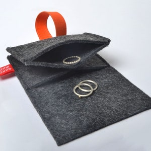 以下が含まれることがあります： 赤い「HAND MADE」のラベルとオレンジ色のゴムループが付いたグレーのフェルト製のリングホルダー。ホルダーが開いており、中に銀色のリングが2つ入っています。