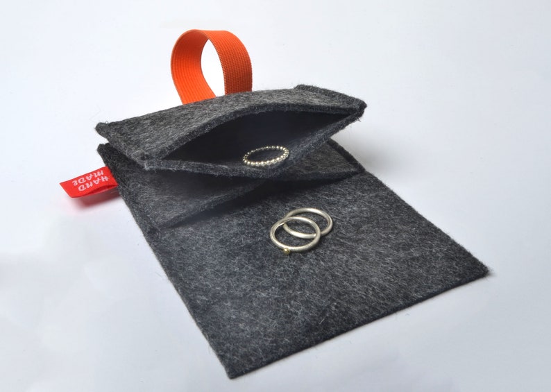 以下が含まれることがあります： 赤い「HAND MADE」のラベルとオレンジ色のゴムループが付いたグレーのフェルト製のリングホルダー。ホルダーが開いており、中に銀色のリングが2つ入っています。