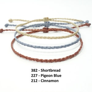May include: Three braided bracelets in different colours: a light brown, a light blue, and a reddish brown. The bracelets are arranged in a circle, with the light brown bracelet on top. The text below the bracelets reads: "382 - Shortbread, 227 - Pigeon Blue, 212 - Cinnamon."