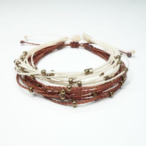 Op de afbeelding: Een meerstrengige armband met bruine en witte draden en gouden kralen.