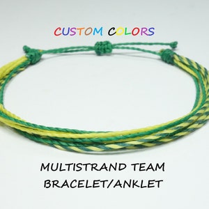 Può includere: Un braccialetto o una cavigliera multi-filo realizzata con corda verde e gialla. Il testo "CUSTOM COLORS" è sopra il braccialetto e il testo "MULTISTRAND TEAM BRACELET/ANKLET" è sotto il braccialetto.