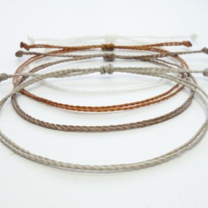 May include: Four braided cord bracelets in different shades of brown and white. The bracelets are layered on top of each other.