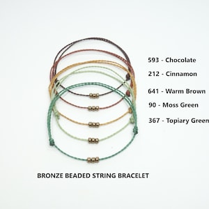 Könnte beinhalten: Fünf verschiedene farbige Armbänder aus Schnur mit Bronzeperlen. Die Farben sind Schokolade, Zimt, warmes Braun, Moosgrün und Topiary-Grün. Der Text "BRONZE-PERLEN-SCHNUR-ARMBAND" befindet sich am unteren Rand des Bildes.