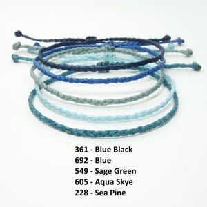 K&ouml;nnte beinhalten: Ein Stapel von sechs geflochtenen Armb&auml;ndern in Blaut&ouml;nen, Gr&uuml;n und Schwarz. Die Armb&auml;nder sind mit ihren Farbnamen und den entsprechenden Nummern beschriftet: 361 - Blau Schwarz, 692 - Blau, 549 - Salbei Gr&uuml;n, 605 - Aqua Skye, 228 - Meereskiefer.