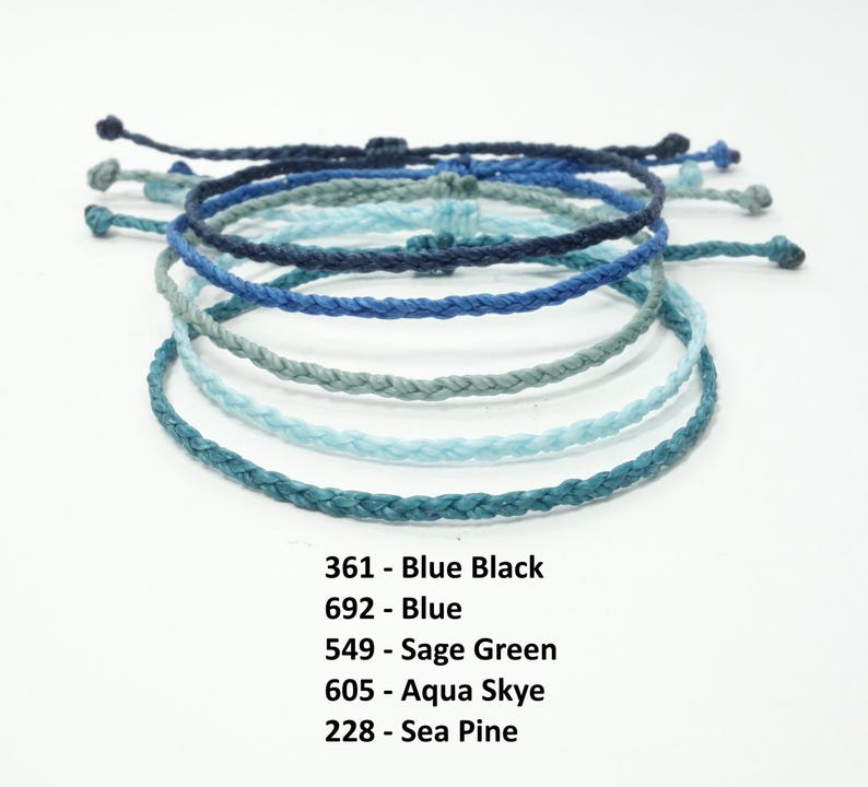 K&ouml;nnte beinhalten: Ein Stapel von sechs geflochtenen Armb&auml;ndern in Blaut&ouml;nen, Gr&uuml;n und Schwarz. Die Armb&auml;nder sind mit ihren Farbnamen und den entsprechenden Nummern beschriftet: 361 - Blau Schwarz, 692 - Blau, 549 - Salbei Gr&uuml;n, 605 - Aqua Skye, 228 - Meereskiefer.