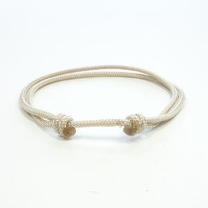 Peut inclure: Un bracelet beige clair en cordon tressé. Le bracelet comporte deux nœuds de chaque côté d'une section centrale en cordon, créant un design réglable. La couleur neutre et le design simple le rendent adapté au quotidien.