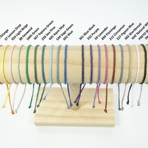 May include: A selection of braided bracelets in a range of colours, such as red, orange, green, blue, and purple. Each bracelet is displayed on a wooden stand, with colour names above. The bracelets are approximately 0.6 cm in diameter.