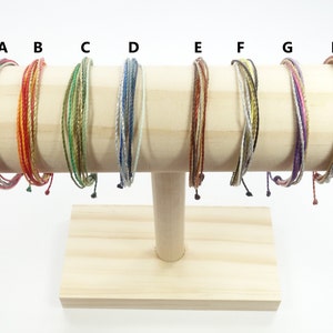 Könnte beinhalten: Acht bunte Armbänder, die auf einem Holzständer ausgestellt sind. Die Armbänder bestehen aus geflochtenem Garn in verschiedenen Farben, darunter Rot, Orange, Gelb, Grün, Blau, Lila und Braun.