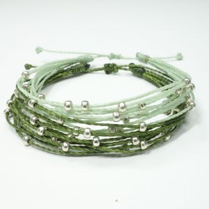 May include: A multi-strand bracelet with a gradient of green and light green colors. The bracelet is adorned with small silver beads.