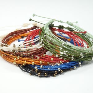 K&ouml;nnte beinhalten: Ein Stapel bunter Armb&auml;nder mit silbernen Perlen. Die Armb&auml;nder bestehen aus d&uuml;nnen, geflochtenen Schn&uuml;ren in verschiedenen Farben, darunter Rot, Blau, Gr&uuml;n und Braun.