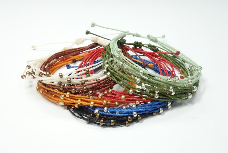 K&ouml;nnte beinhalten: Ein Stapel bunter Armb&auml;nder mit silbernen Perlen. Die Armb&auml;nder bestehen aus d&uuml;nnen, geflochtenen Schn&uuml;ren in verschiedenen Farben, darunter Rot, Blau, Gr&uuml;n und Braun.
