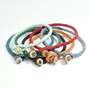 K&ouml;nnte beinhalten: Ein Set aus acht bunten geflochtenen Armb&auml;ndern mit Holz-Knopfverschl&uuml;ssen. Die Armb&auml;nder sind in verschiedenen Farben, darunter Blau, Gr&uuml;n, Rot und Orange.