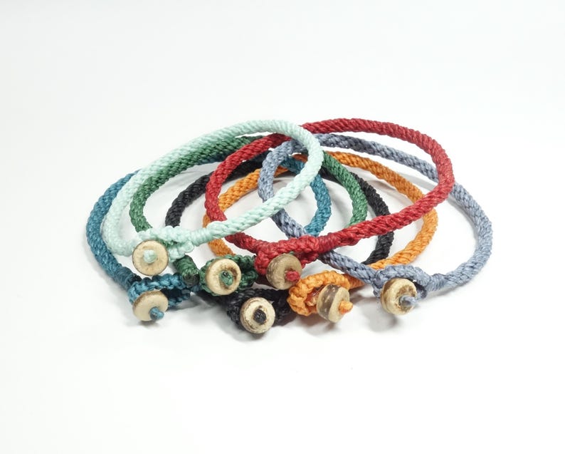 K&ouml;nnte beinhalten: Ein Set aus acht bunten geflochtenen Armb&auml;ndern mit Holz-Knopfverschl&uuml;ssen. Die Armb&auml;nder sind in verschiedenen Farben, darunter Blau, Gr&uuml;n, Rot und Orange.