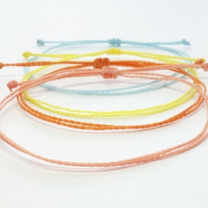 Può includere: Set di quattro braccialetti dell'amicizia colorati in tonalità blu chiaro, giallo, arancione e rosa. I braccialetti sono realizzati con un cordoncino sottile intrecciato e sono legati con un semplice nodo.