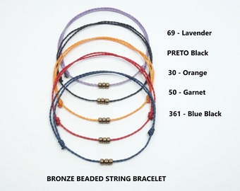 May include: Five colorful string bracelets with bronze beads. The bracelets are arranged in a stack, with the lavender bracelet on top and the blue black bracelet on the bottom. The text "BRONZE BEADED STRING BRACELET" is at the bottom of the image. The text "69 - Lavender", "PRETO Black", "30 - Orange", "50 - Garnet", and "361 - Blue Black" is on the right side of the image.