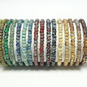 Könnte beinhalten: Eine Sammlung von 18 bunten Armbändern aus kleinen Glasperlen. Die Armbänder sind in einer Reihe angeordnet, wobei die Farben von Grün nach Braun übergehen.