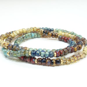 Op de afbeelding: Een meerstrengs armband gemaakt van kleine, kleurrijke glazen kralen. De kralen zijn in tinten bruin, goud, blauw en groen. De armband is gelaagd en heeft een delicate, bohemian stijl.