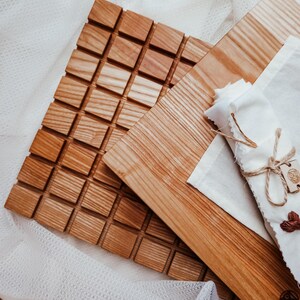 Könnte beinhalten: Holzservierbretter und ein quadratischer Untersetzer mit einem strukturierten, würfelartigen Muster. Eine weiße Stoffserviette ist mit Bindfaden gebunden. Die Bretter und der Untersetzer sind aus hellbraunem Holz.